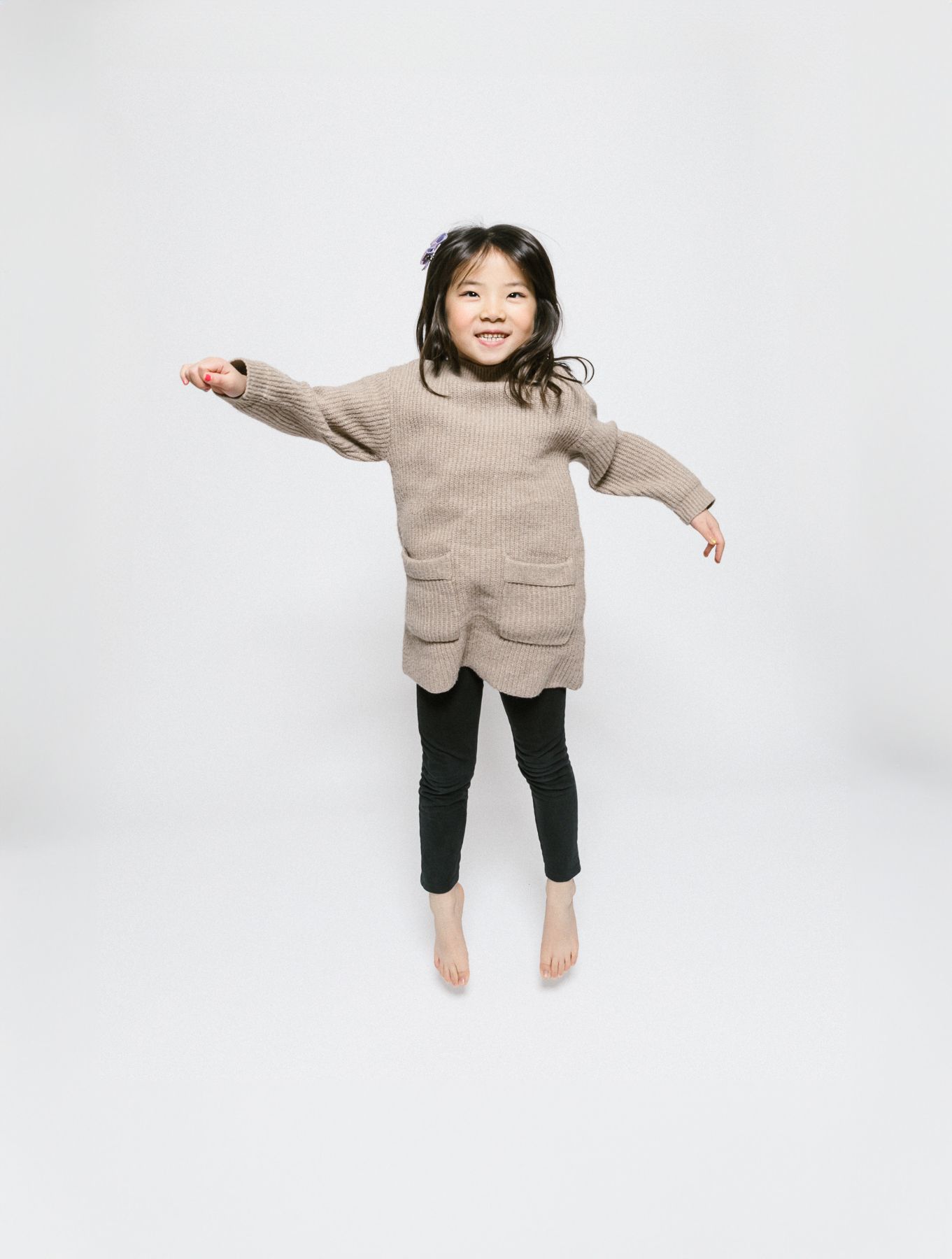 A child in a beige sweater and black leggings jumping joyfully against a white background.