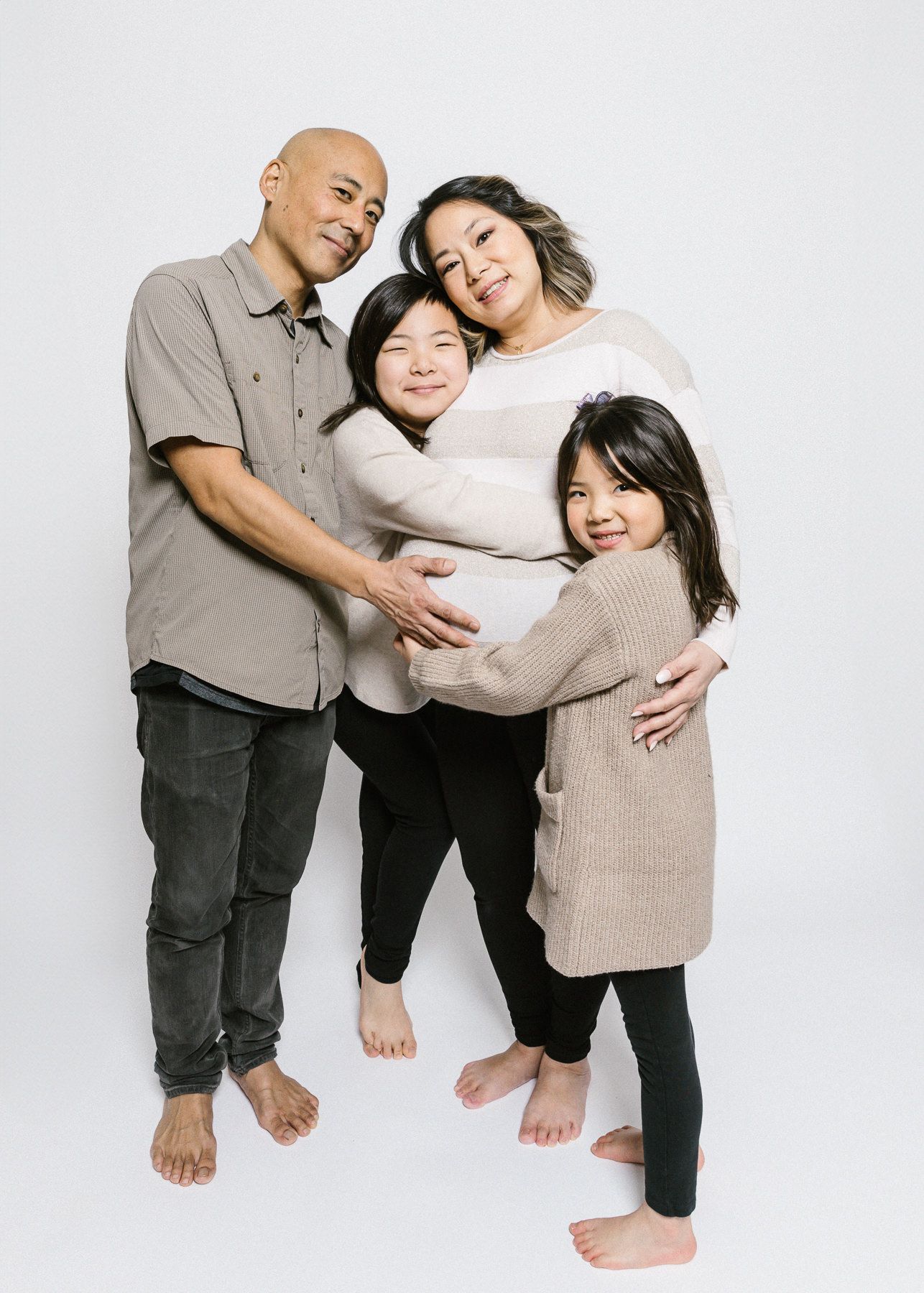 Family of four wearing coordinating neutral colors posing against white background.
