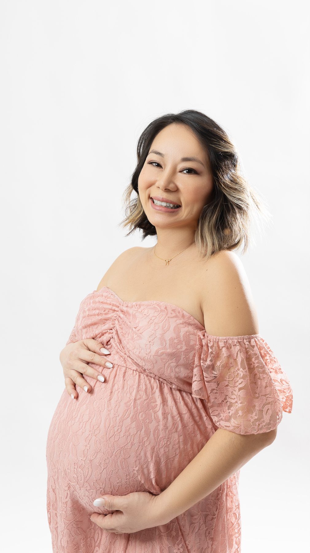 Maternity portrait in pink lace dress against white background.