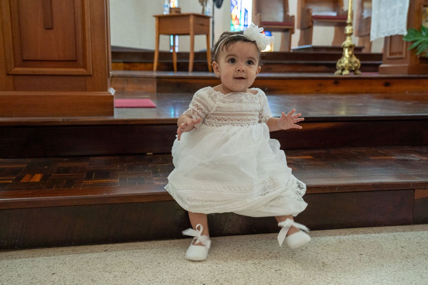Baby in traditional white christening gown sitting on wooden church steps.