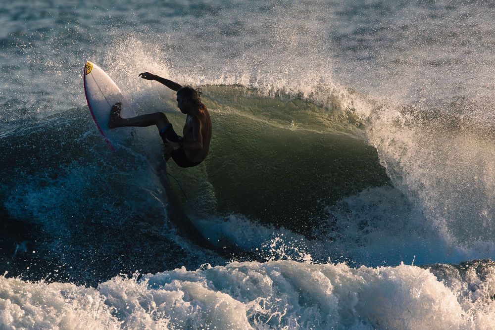 Surfer rides through a powerful ocean wave creating dramatic splashes and spray in the sunlight.