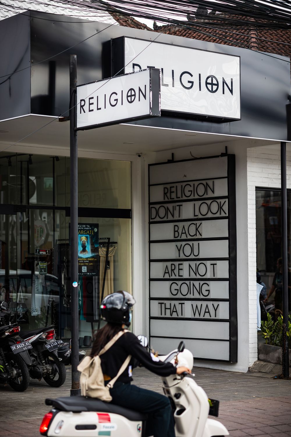 A storefront sign for 'Religion' with text reading 'Look back you are not going that way' and a scooter parked outside.