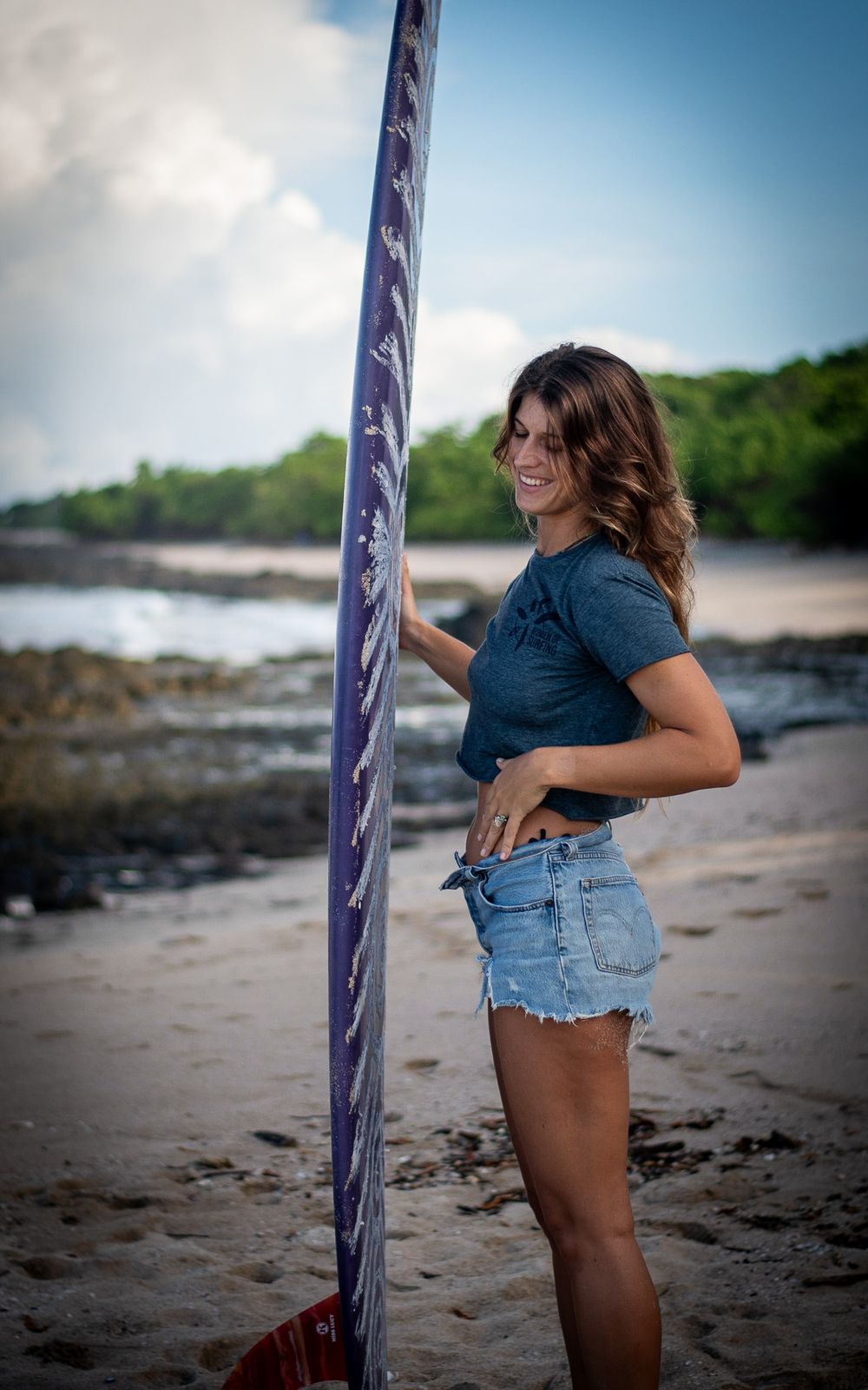 Person in casual beachwear poses with surfboard on rocky coastal shore.