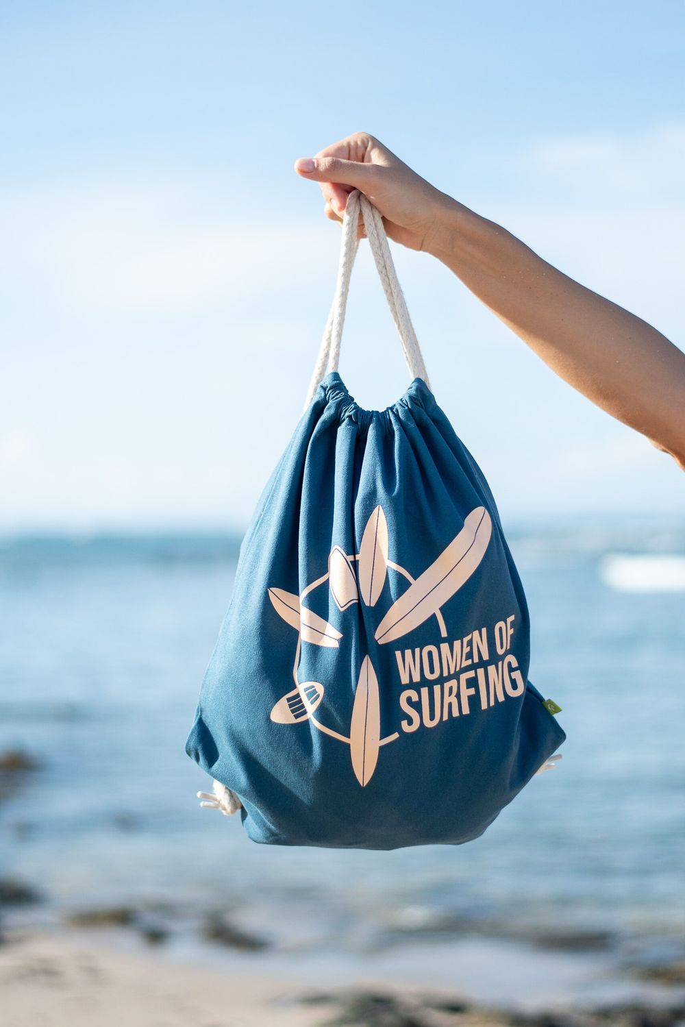 Blue drawstring bag with 'Women of Surfing' logo and surfboard design held up against beach background.