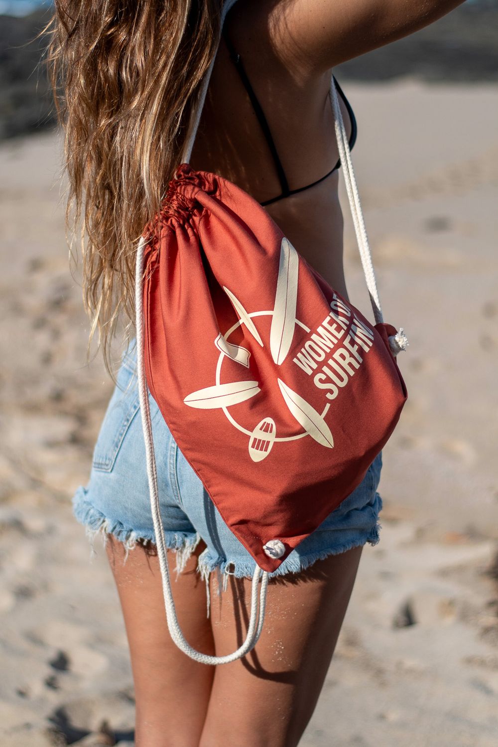 Coral red drawstring bag with surfboard logo worn with denim shorts on beach.