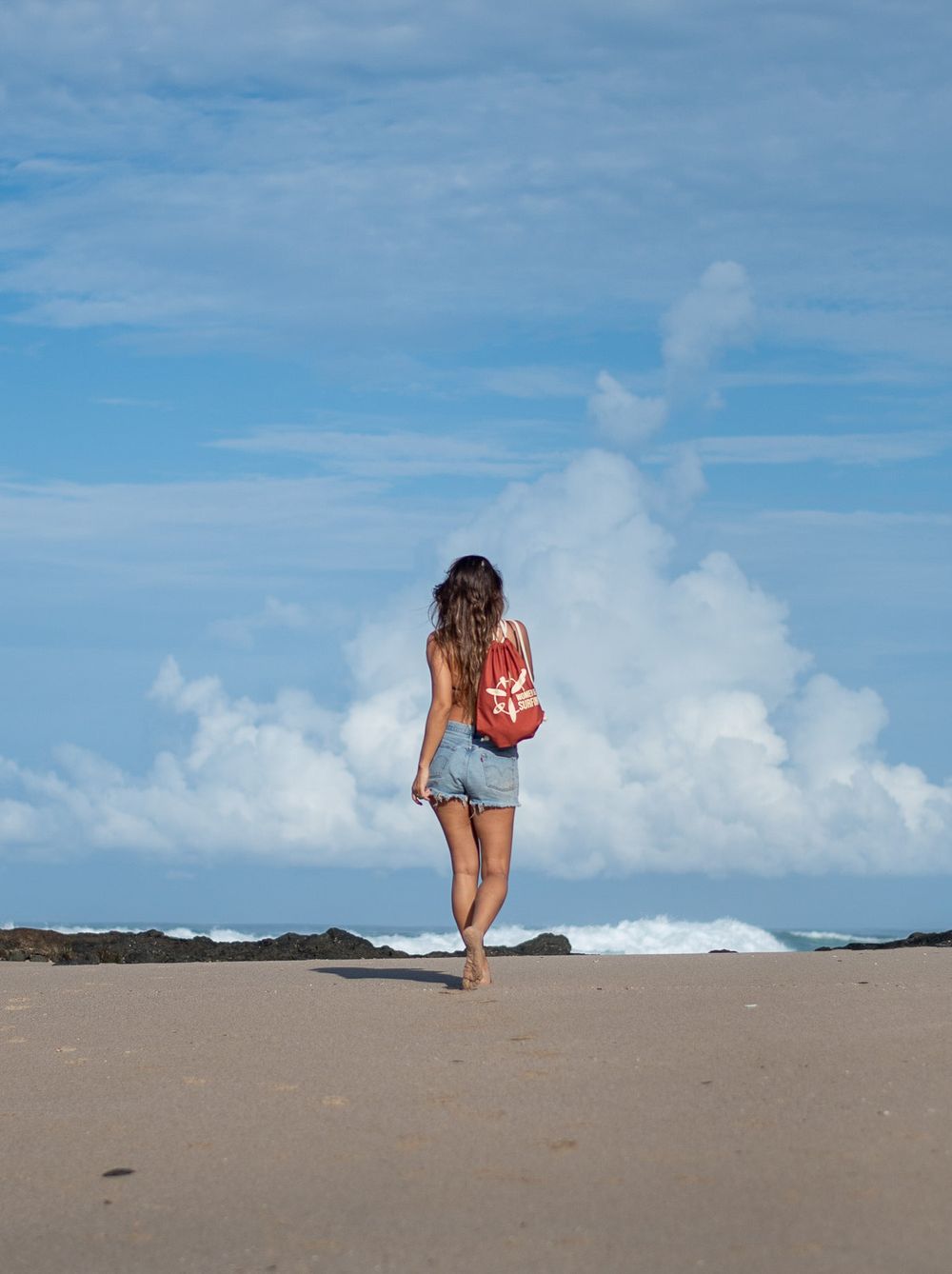 Person walking on sandy beach with red backpack against cloudy ocean horizon.