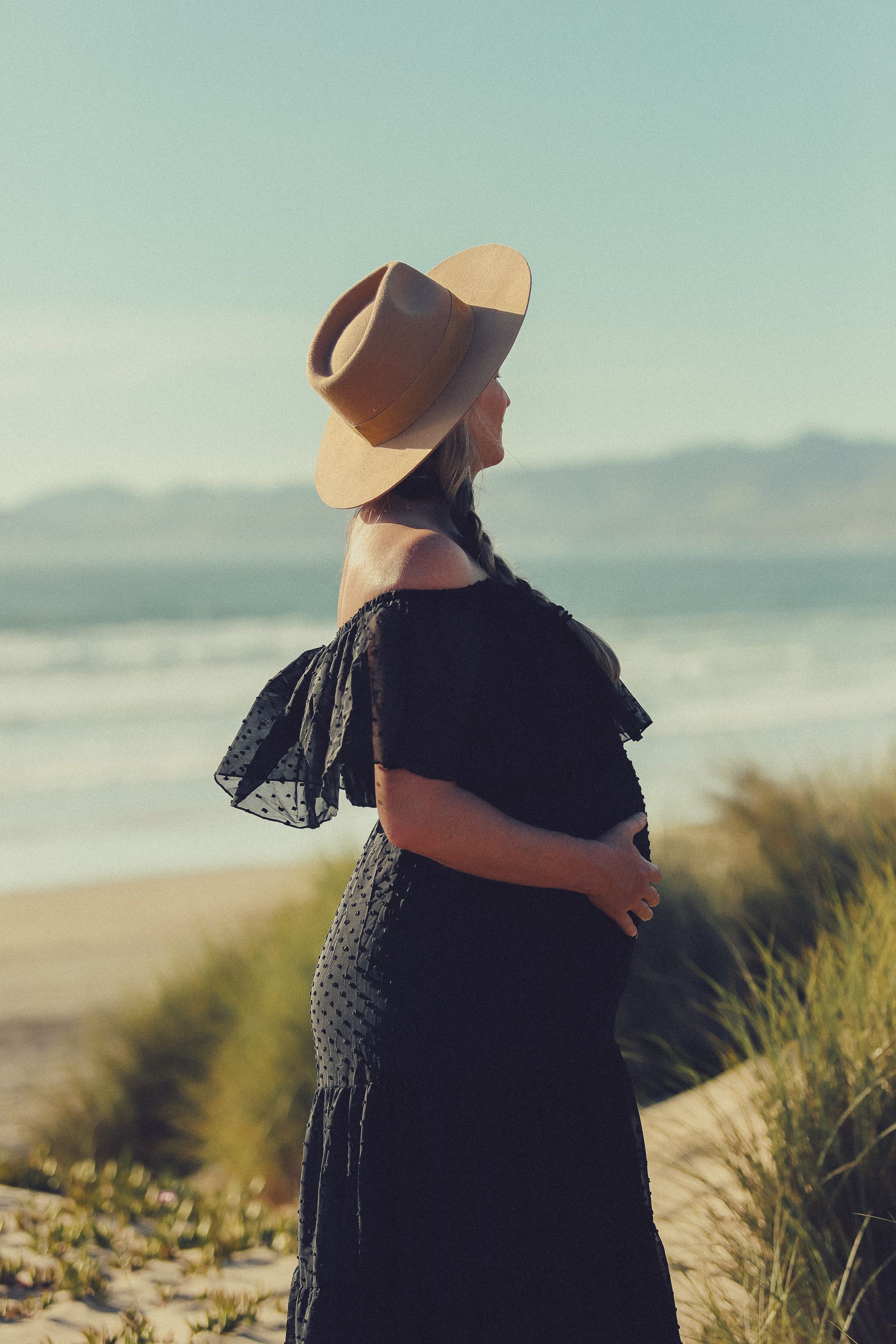 Figure in black dress and hat stands overlooking coastal beach landscape.