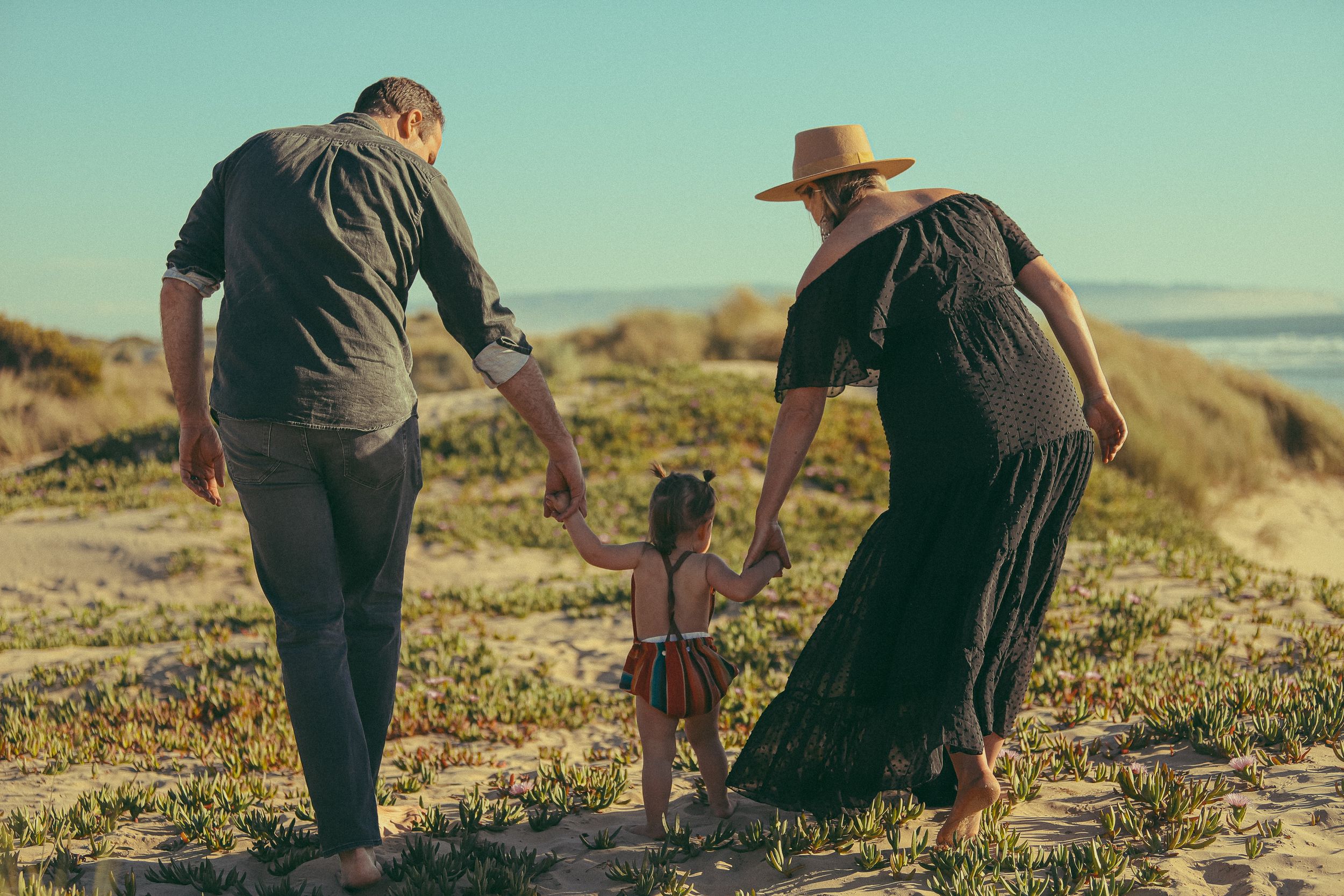Two adults helping a small child walk along a scenic hillside path at sunset.