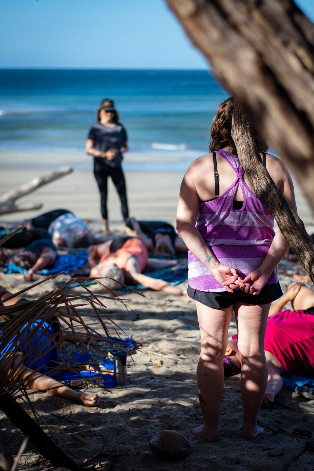 Beachside yoga session participants resting on colorful blankets near driftwood.