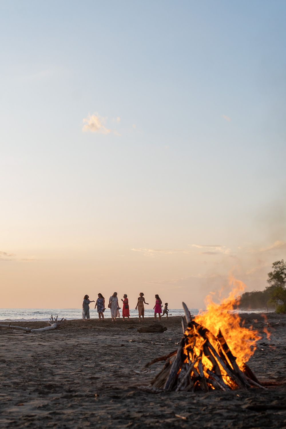 Beach bonfire at sunset with silhouettes of people in background.