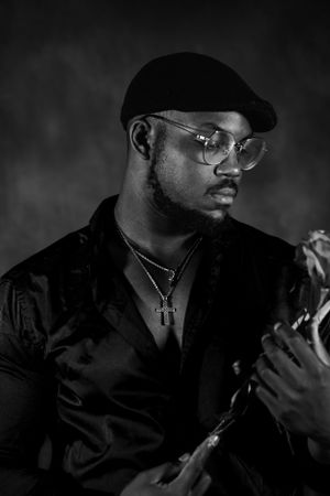 Side profile portrait in low key lighting showing person wearing cap and glasses with cross necklace.