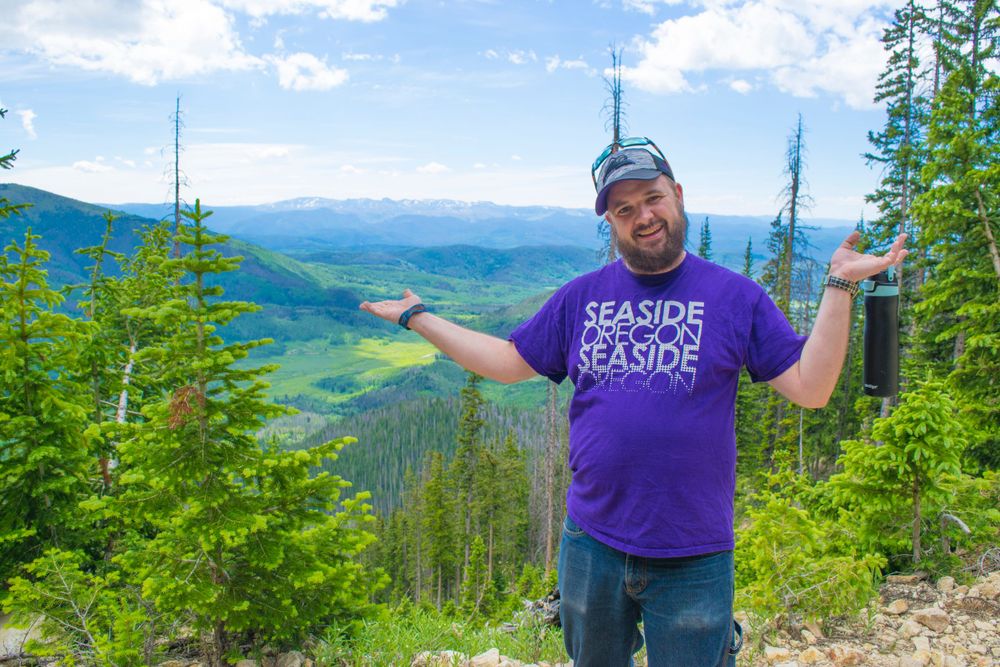 Hiker gestures at scenic mountain overlook while wearing purple t-shirt on sunny day.
