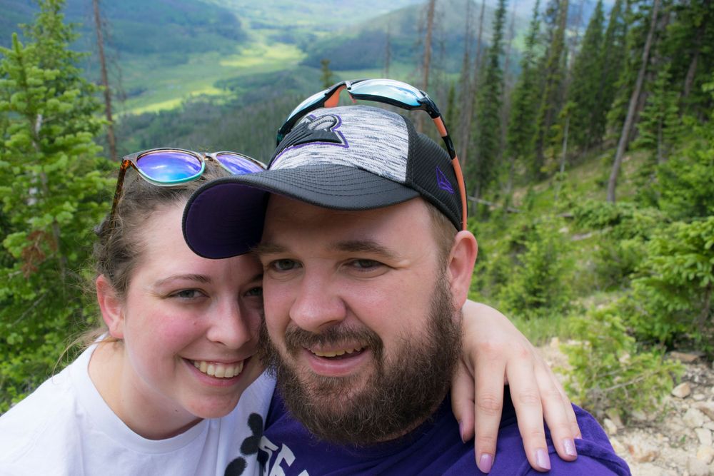 Hiking companions share a moment at scenic mountain overlook with valley views behind them.