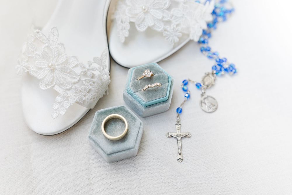 Wedding details with white lace shoes, blue ring box, and rosary on light background.