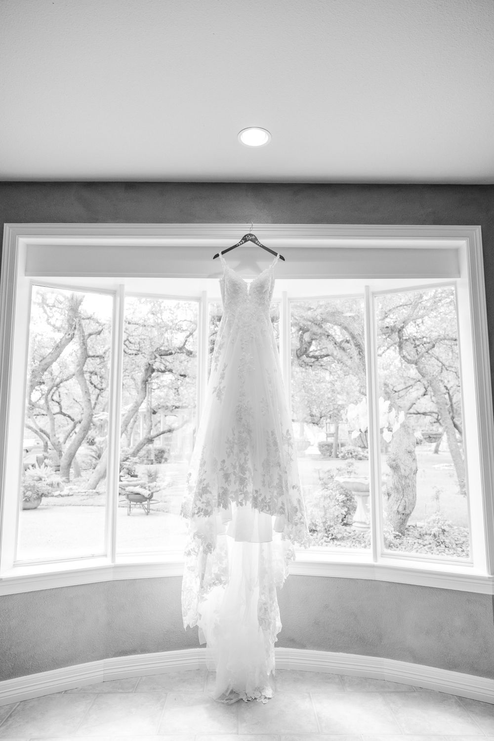 Wedding dress hanging in bay window with natural light creating dramatic silhouette.