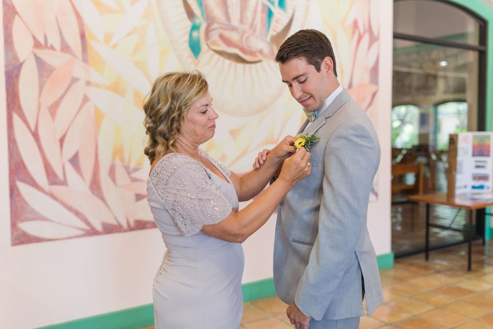 Mother helps adjust boutonniere on groom's light gray suit jacket in front of pastel wall art.