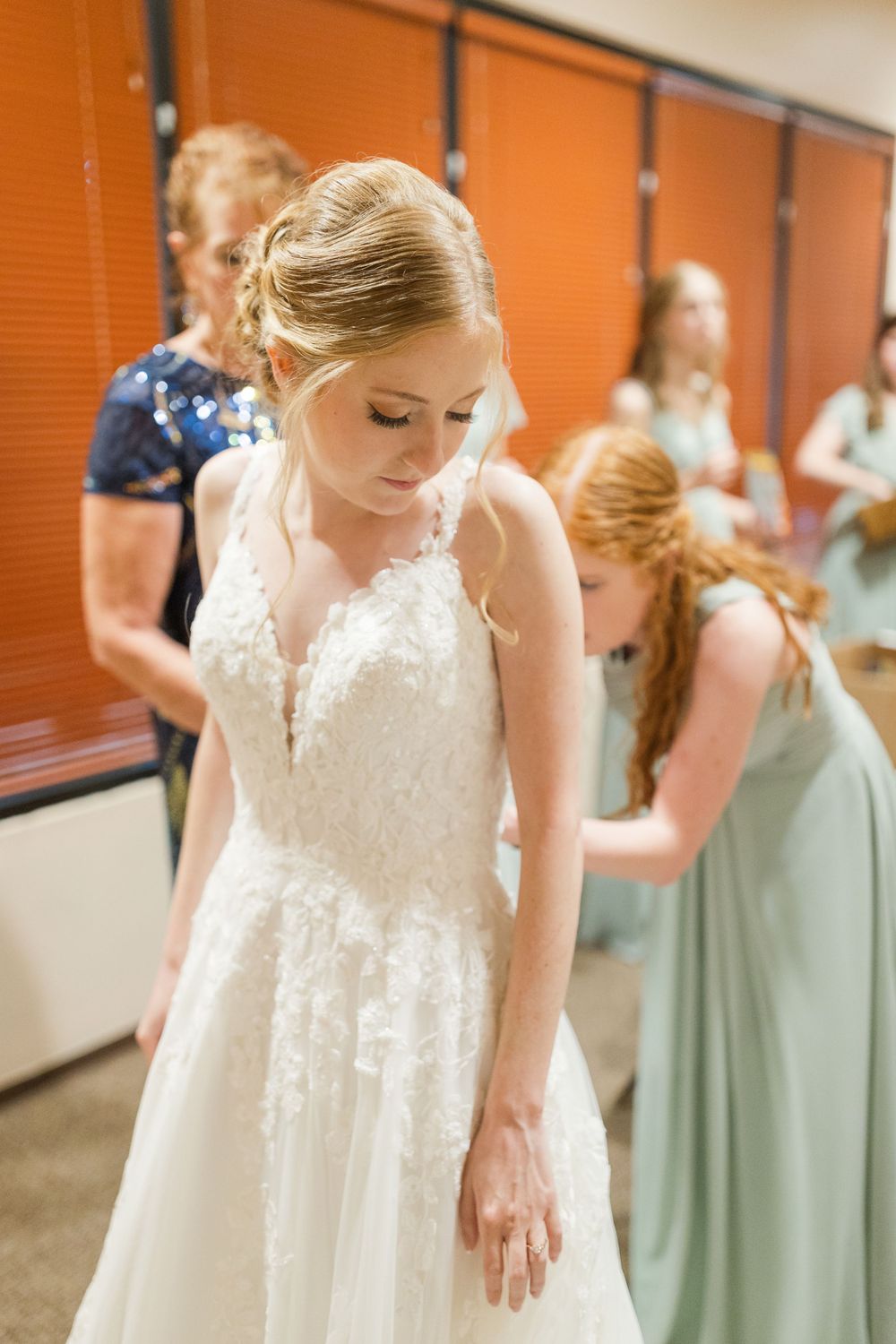 Bride in delicate lace wedding gown stands while getting ready in dressing room.