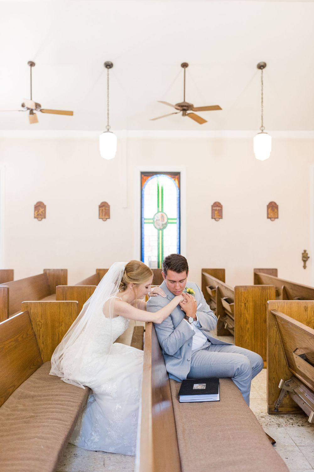 Bride and groom seated in church pew with ceiling fans and stained glass visible above.