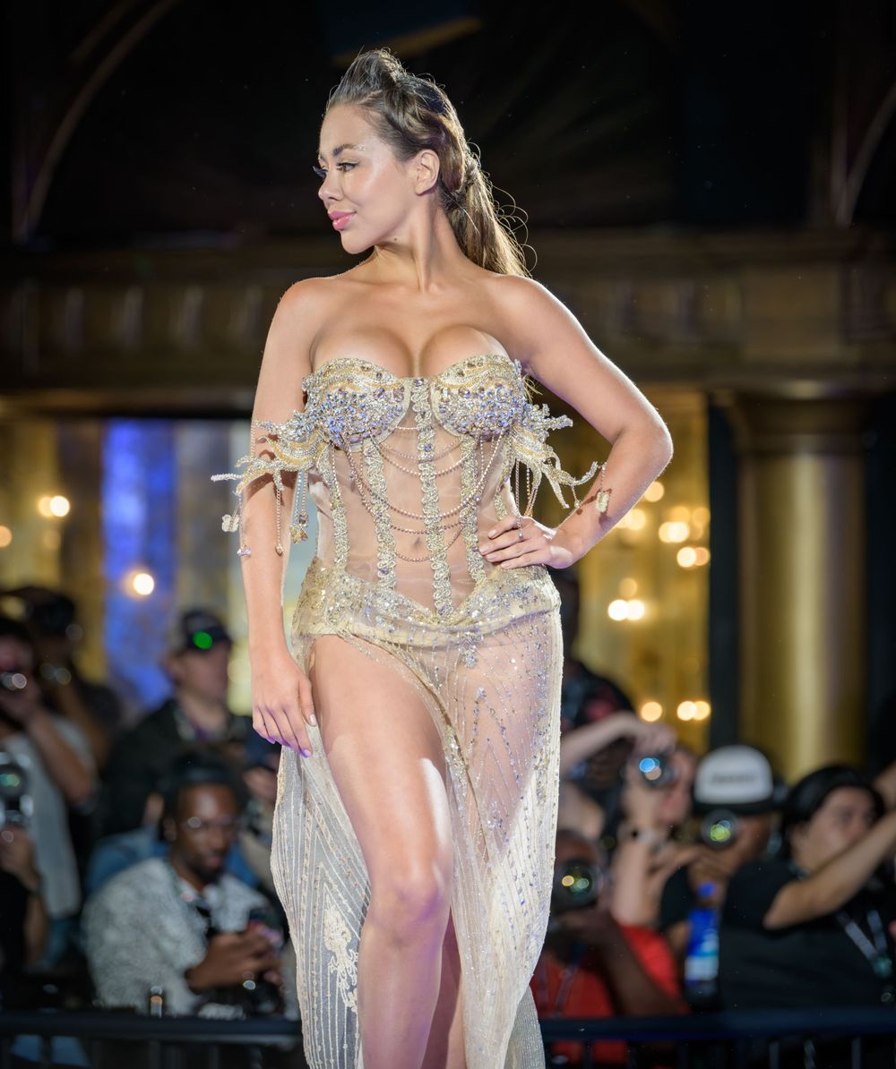 A performer in a glittering sheer dress with a jeweled bodice walks on a runway in front of an audience.