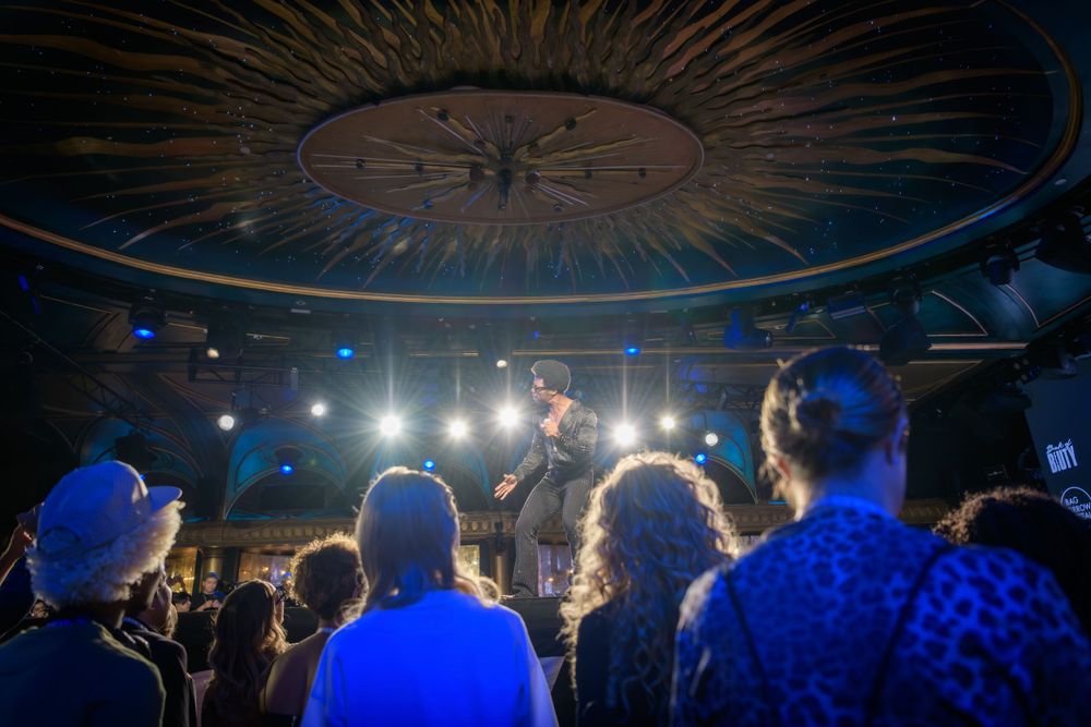 Concert crowd silhouettes illuminated by blue lighting and stage spotlights in domed venue.