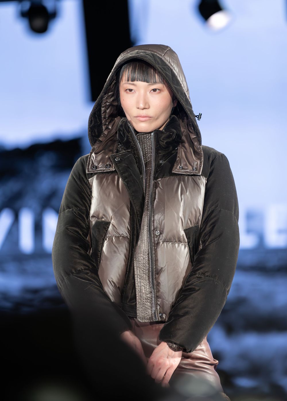 Person wearing a brown puffer vest and black jacket with hood stands against blue winter background.