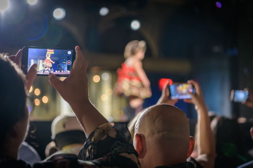 A concert crowd records video on mobile phones while a performer in red stands on stage in a dark venue.