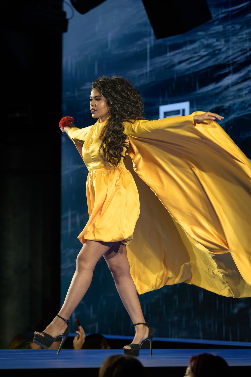 A performer in a flowing yellow dress dances dramatically on stage with fabric billowing behind.