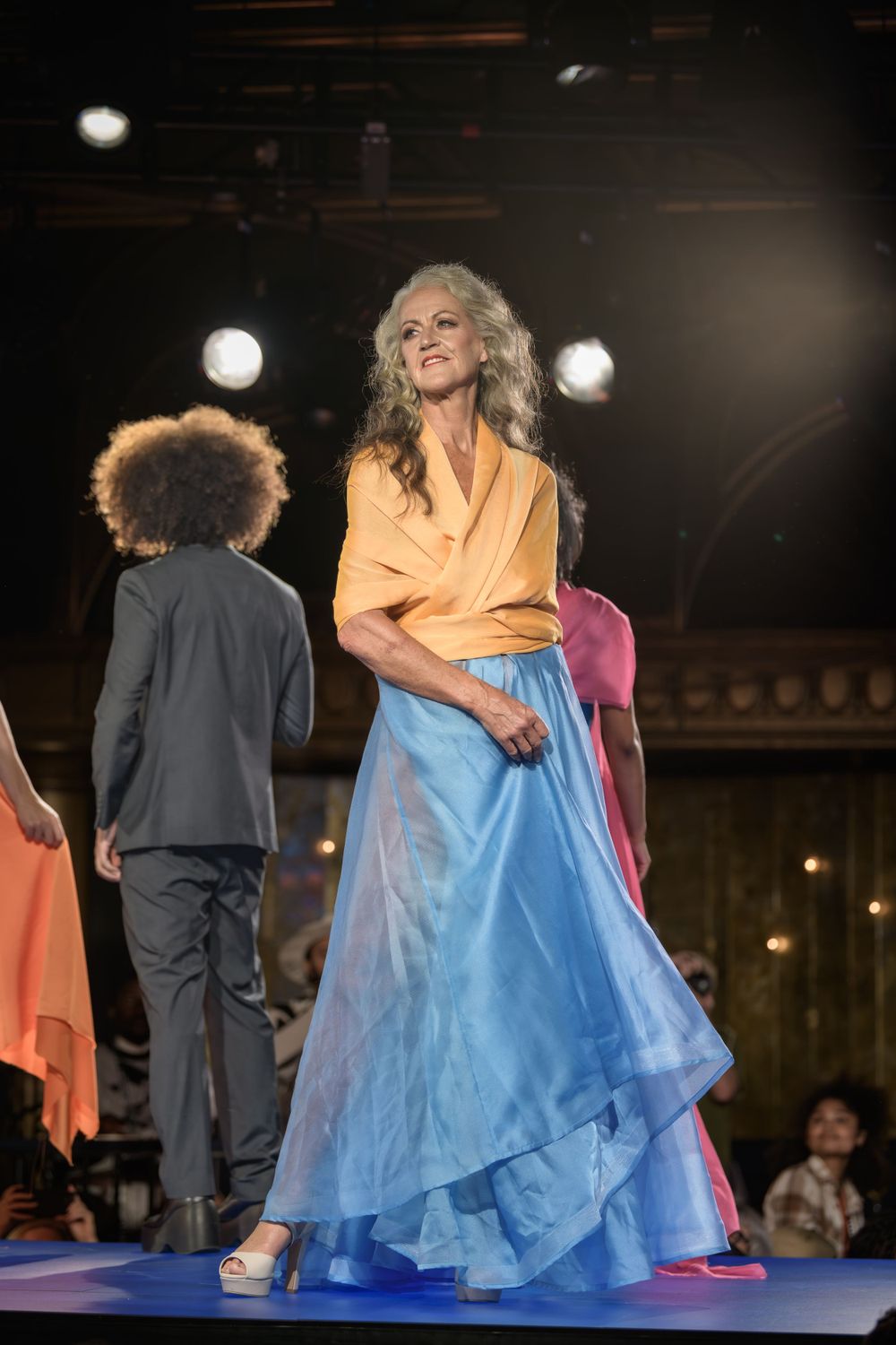 Fashion model walks runway in flowing blue skirt and yellow top during elegant stage performance.