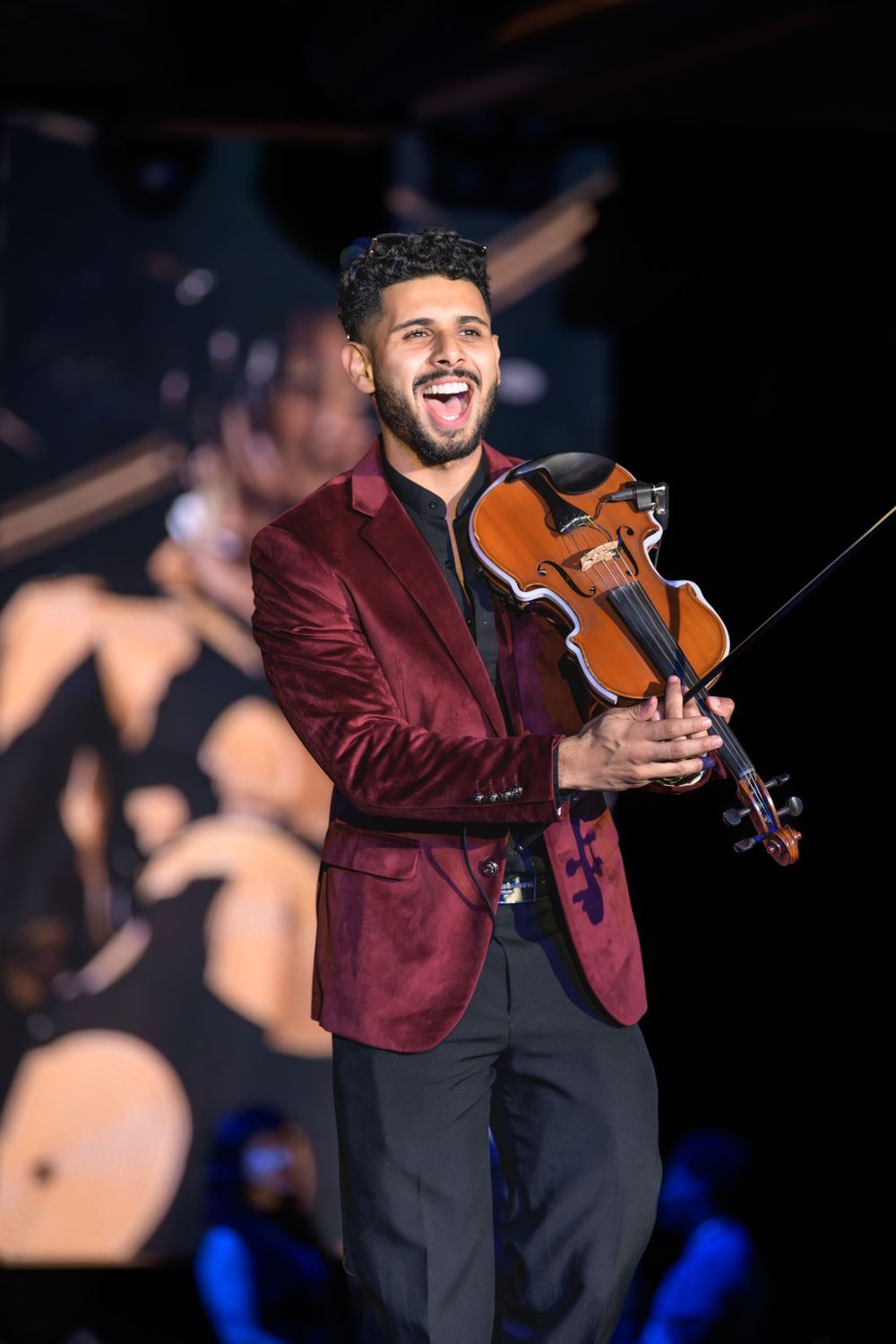 Violinist performs on stage wearing a burgundy blazer with dramatic stage lighting in the background.