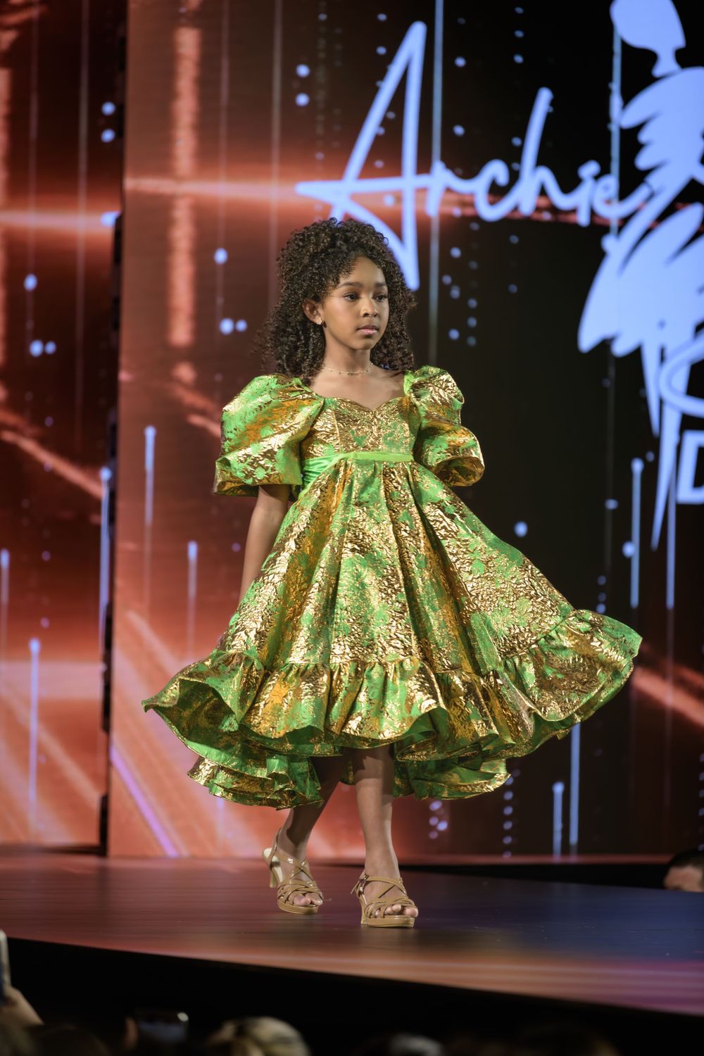 A young model walks the runway in a dramatic lime green ruffled dress with puff sleeves during a fashion show.