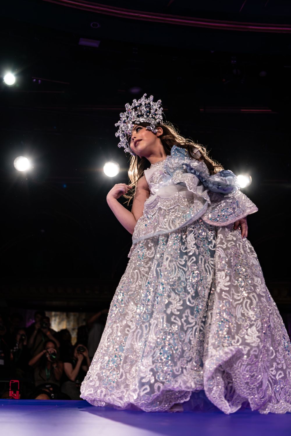A performer in an ornate silver gown and crown walks the runway at a fashion show under dramatic lighting.