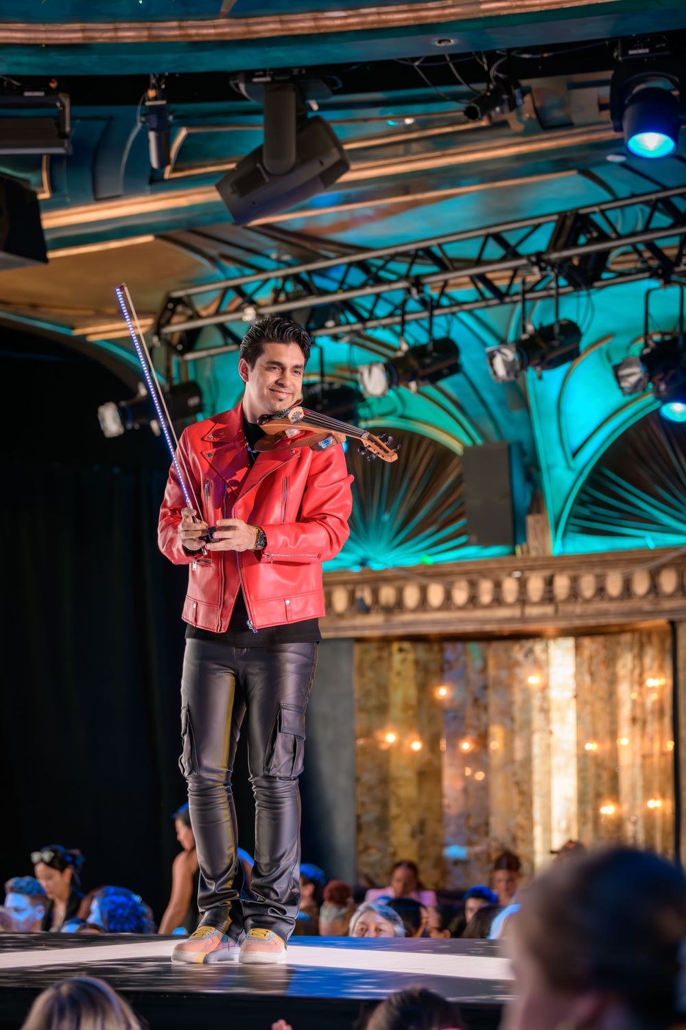 A performer in a bright red jacket plays violin on stage under dramatic turquoise lighting.