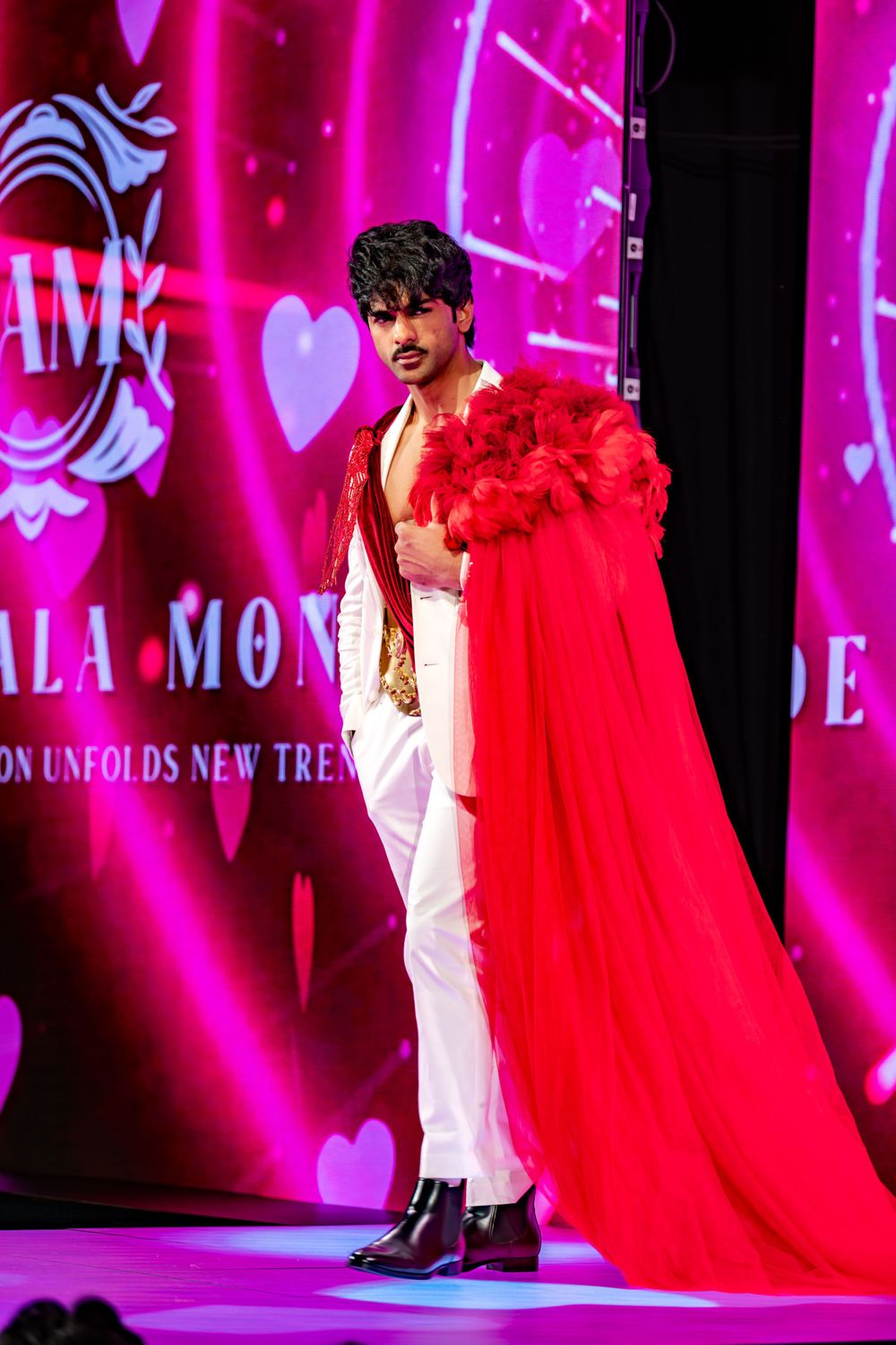 Model walks runway in dramatic red cape and white pants at fashion show with pink neon lighting and heart decorations.