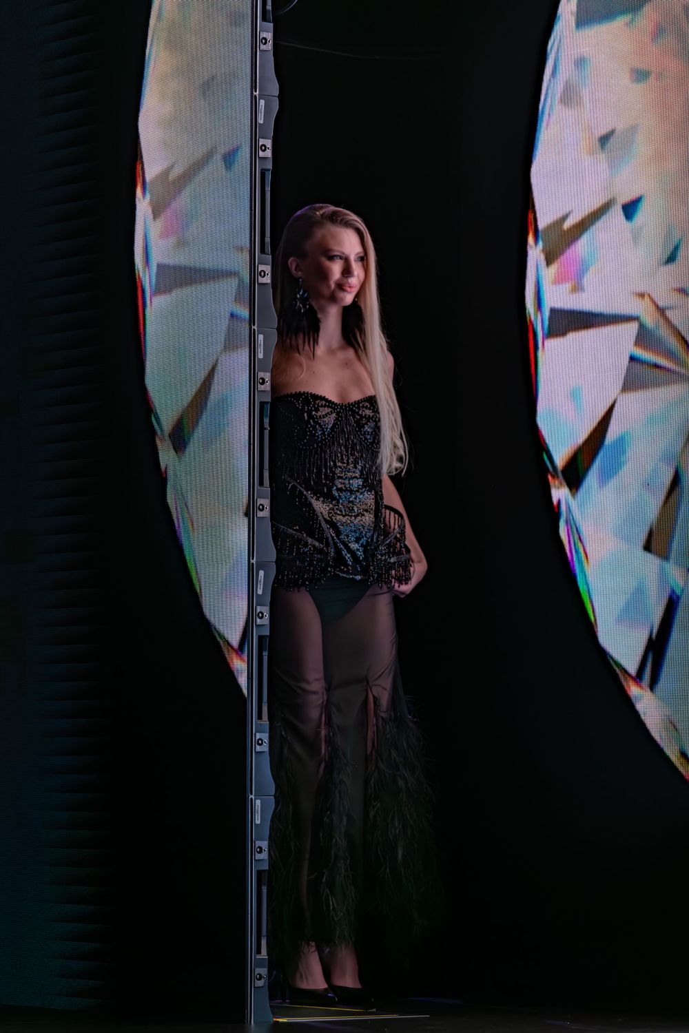 A figure in a dark sequined bodysuit stands between illuminated panels with colorful geometric projections.