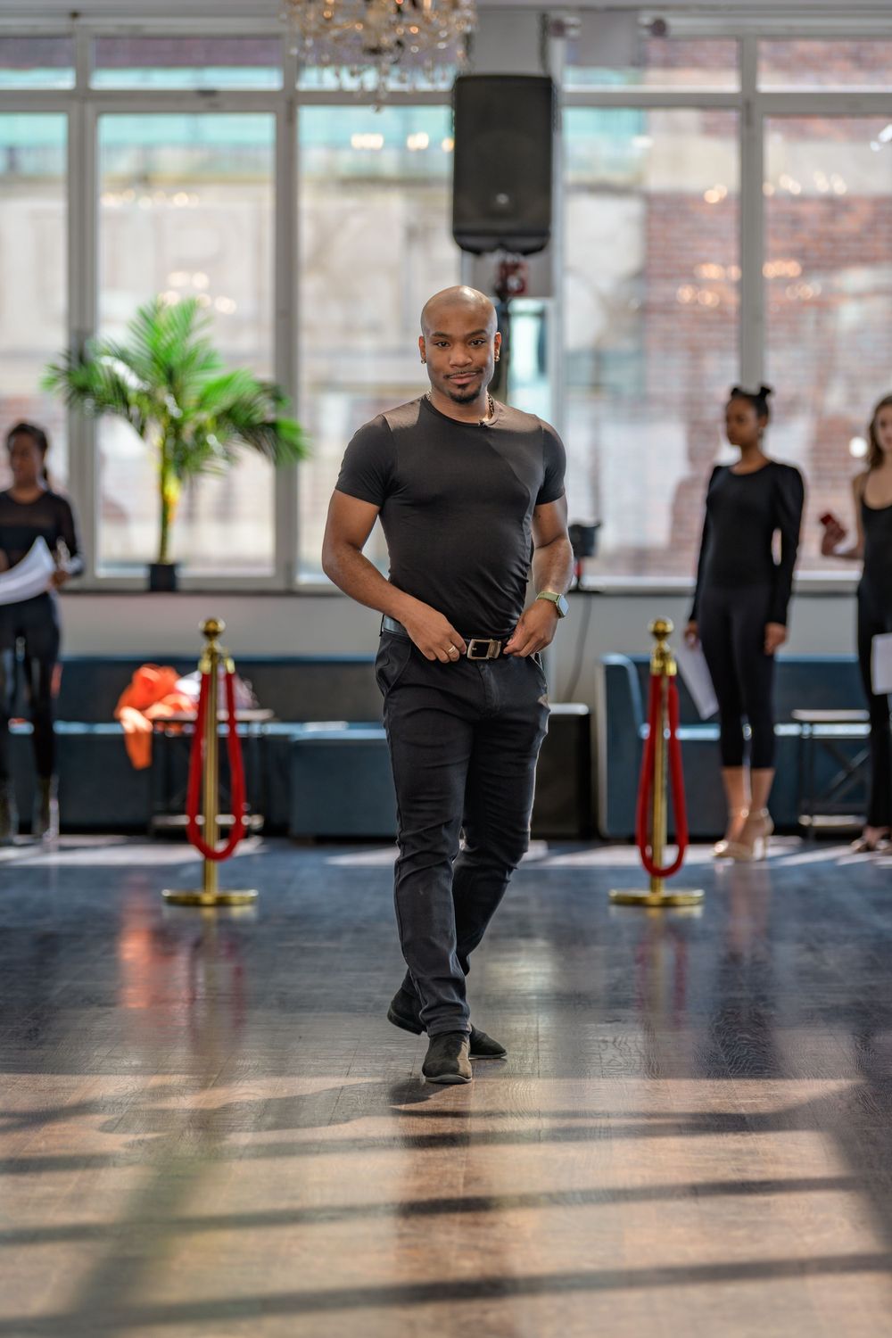 Performer in all-black attire walking across dance studio floor during practice session.