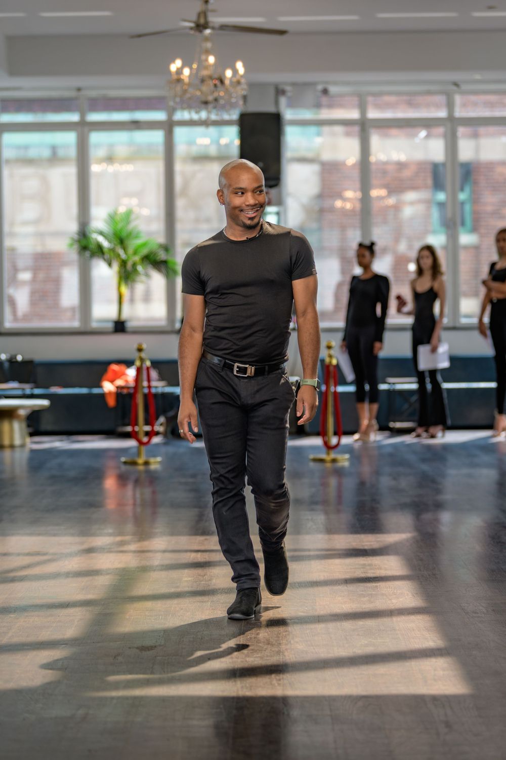 Dance instructor walks across the studio floor in black attire with other dancers practicing in the background.