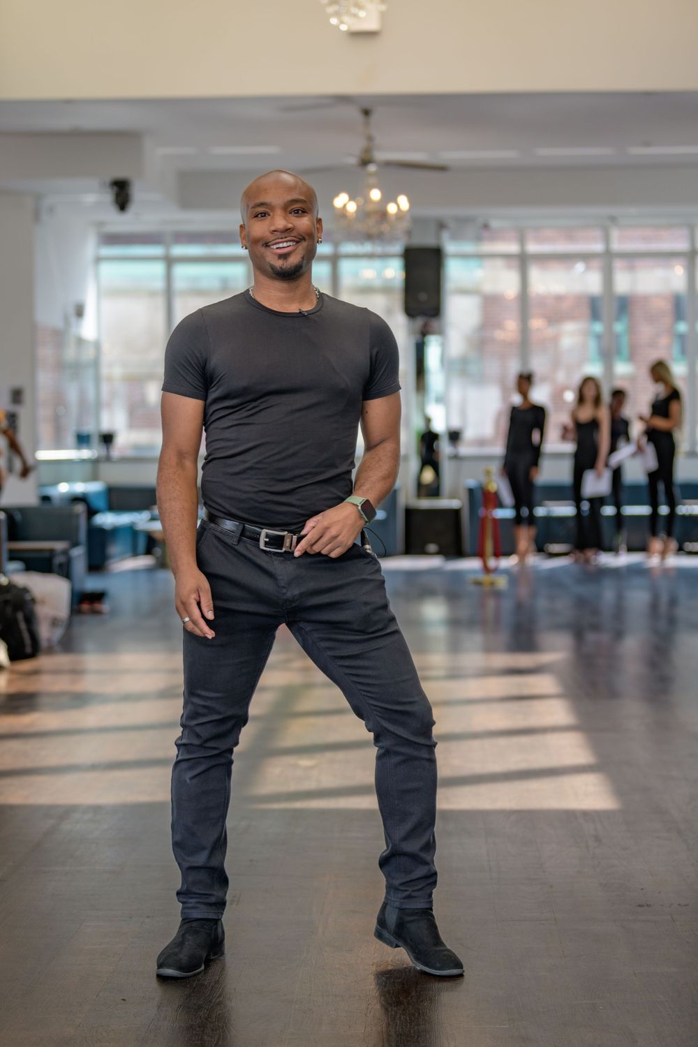 Performer in casual black outfit posing confidently in bright dance studio with chandelier.