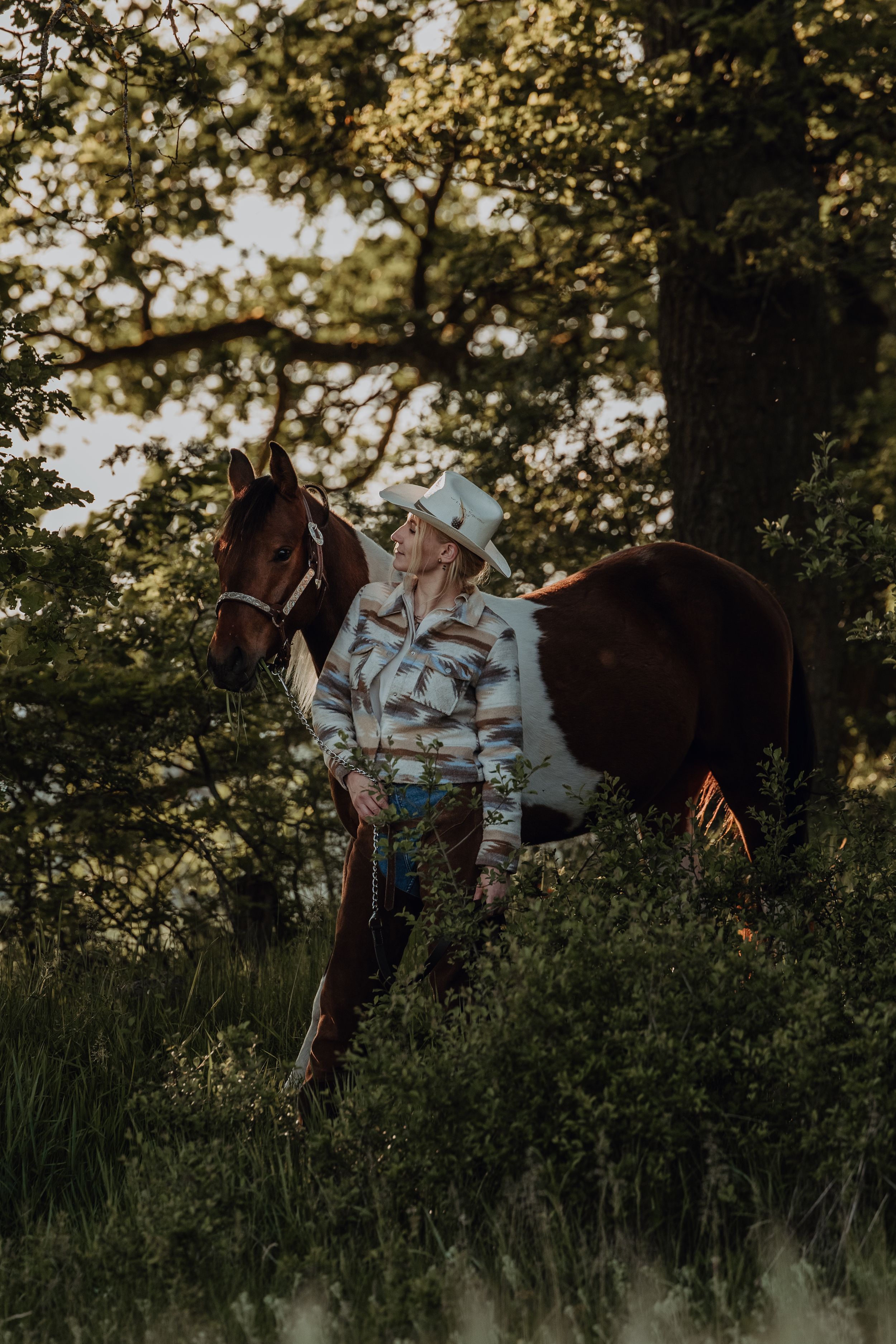 A horse and rider in western gear stand among trees during golden hour.