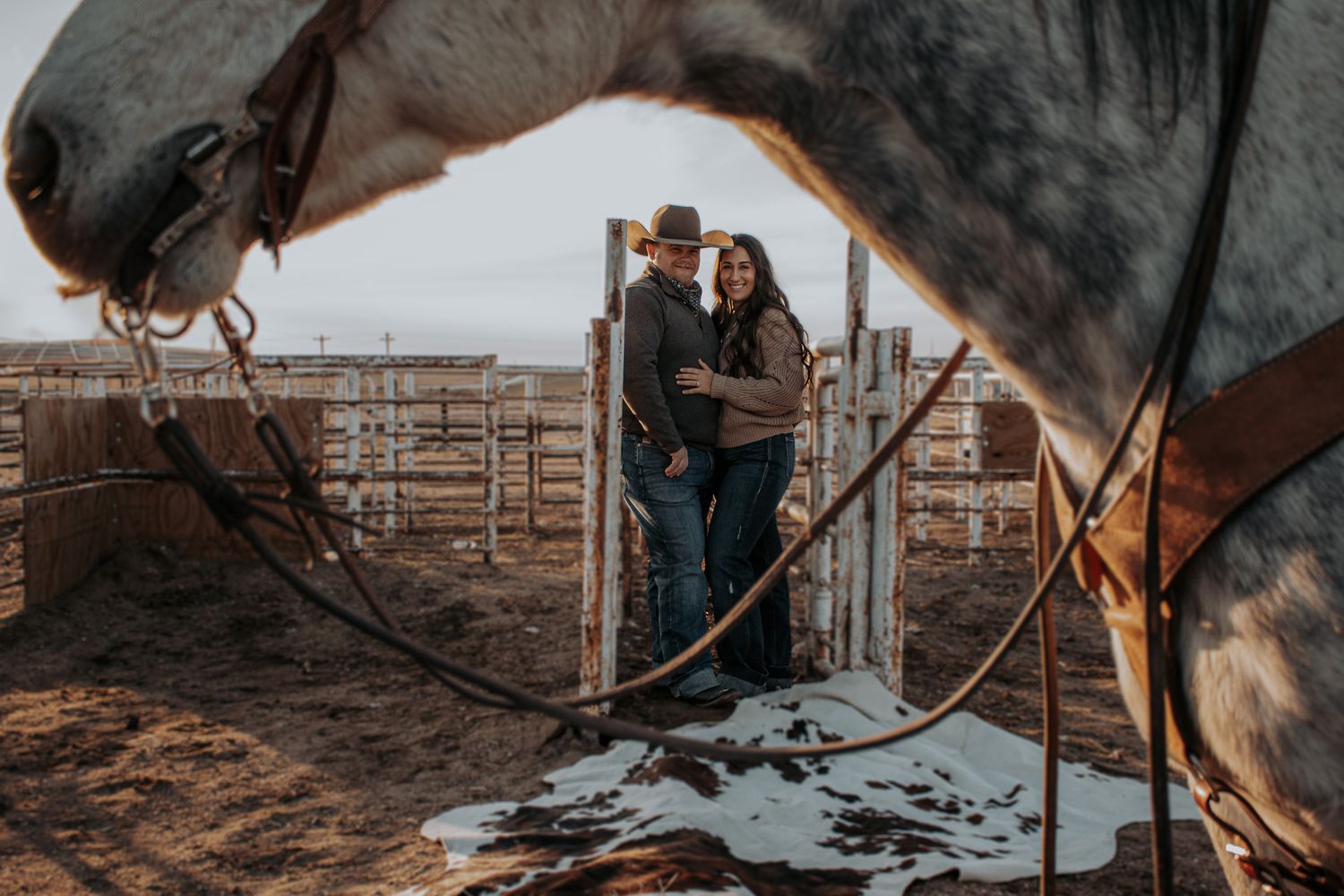 A romantic western scene framed by a horse's neck showing a couple in a rustic corral at sunset.