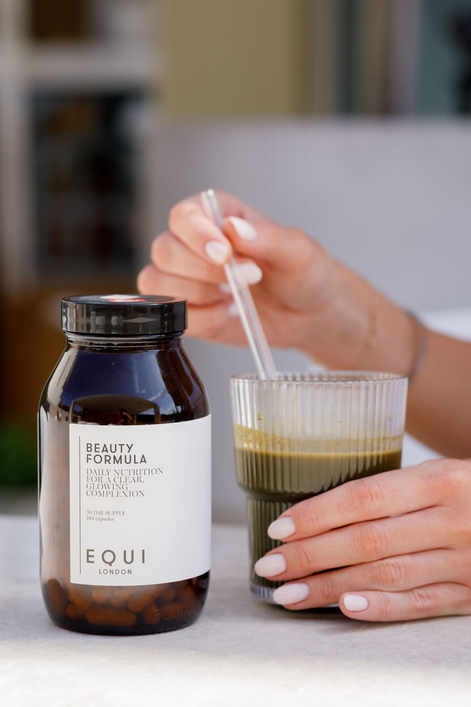Hand holding glass of layered wellness drink next to Equi Beauty Formula supplement bottle on white surface.