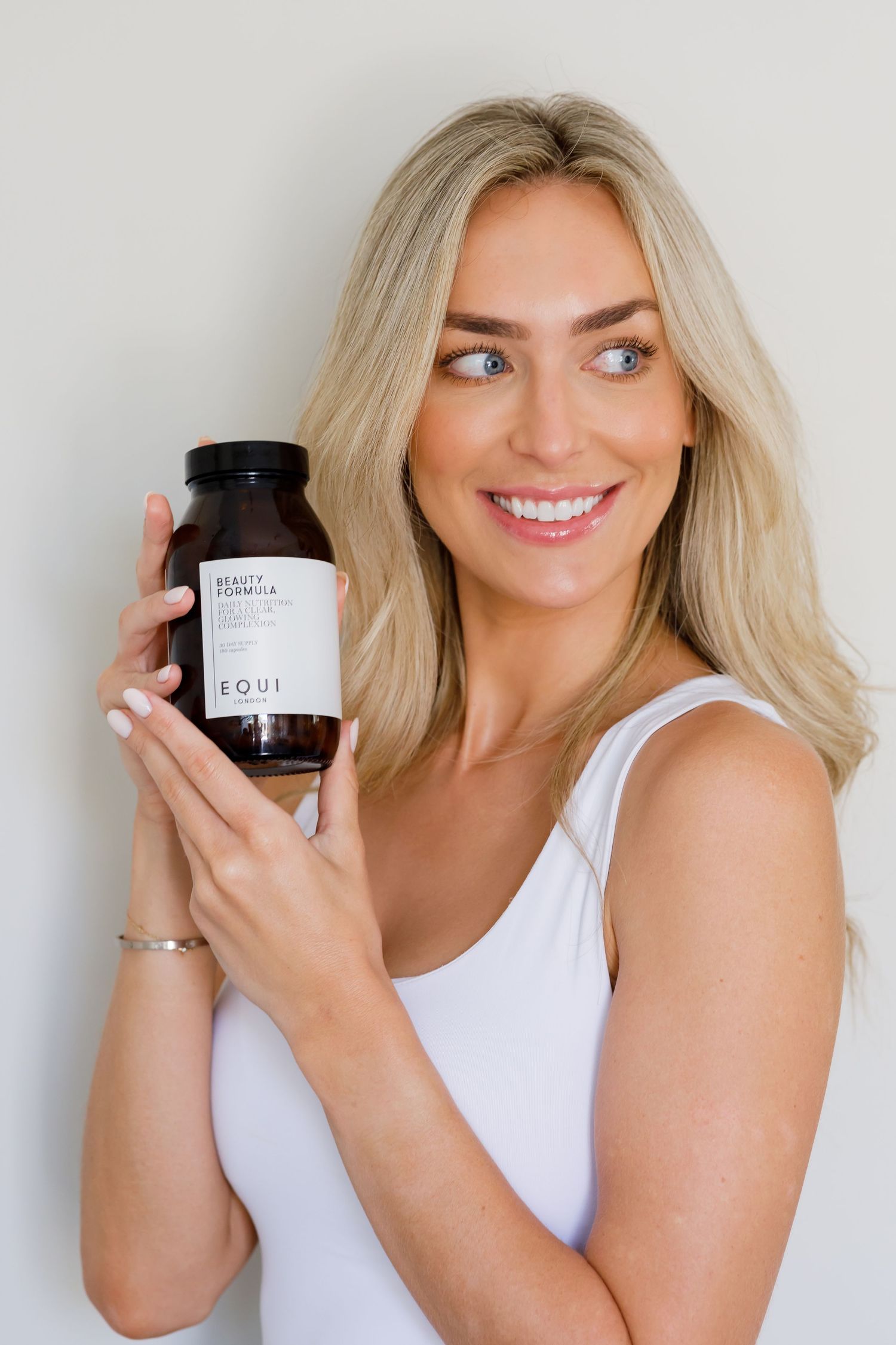 A person wearing a white tank top holding up a dark glass supplement bottle while smiling at the camera.