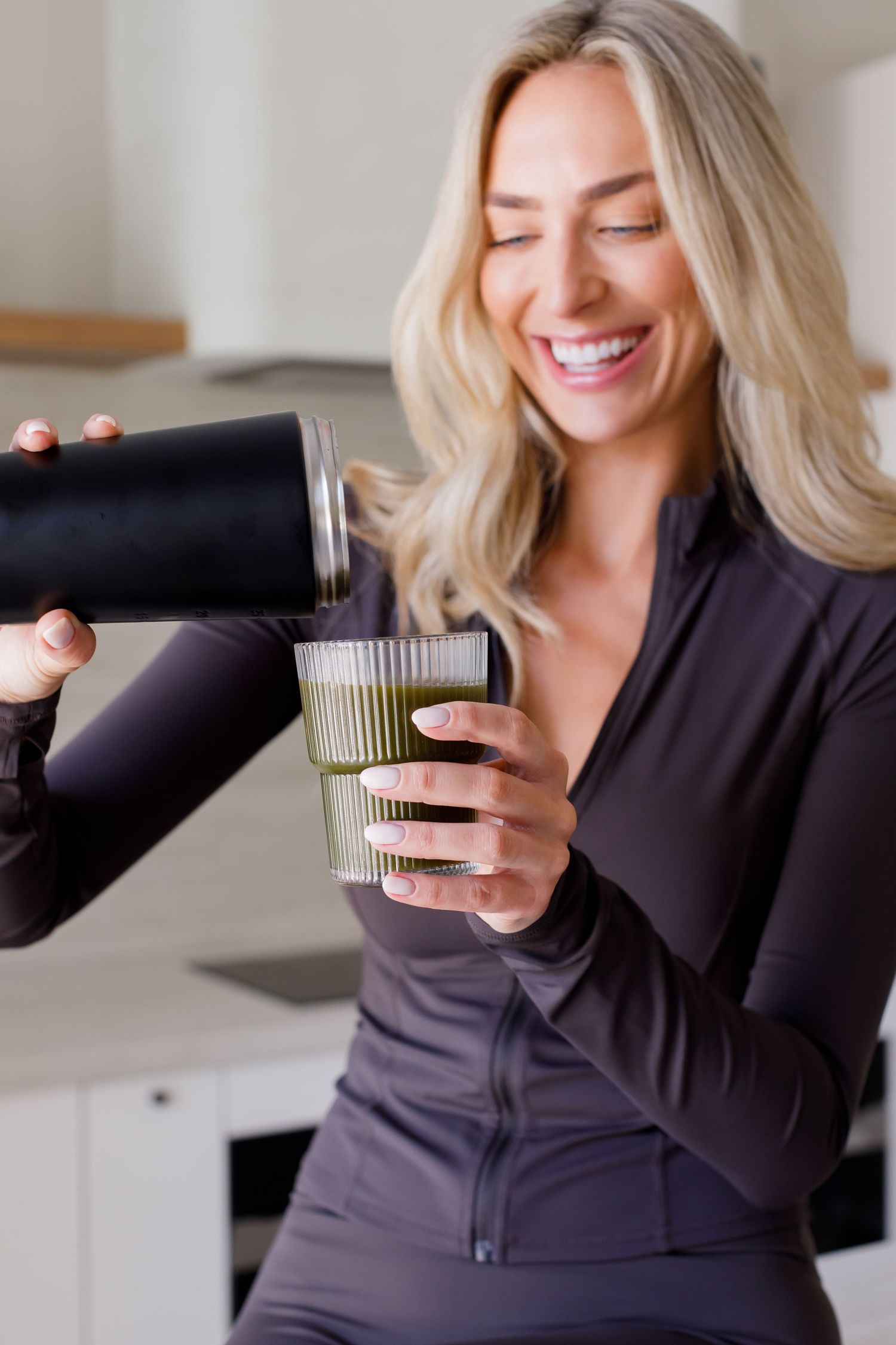 A smiling person in athletic wear pouring a drink from a black container into a reusable cup in a kitchen setting.