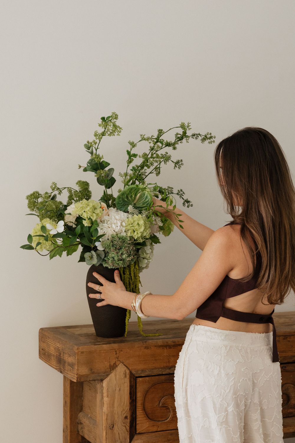 Woman arranging green and white floral bouquet in dark vase on rustic wooden dresser.