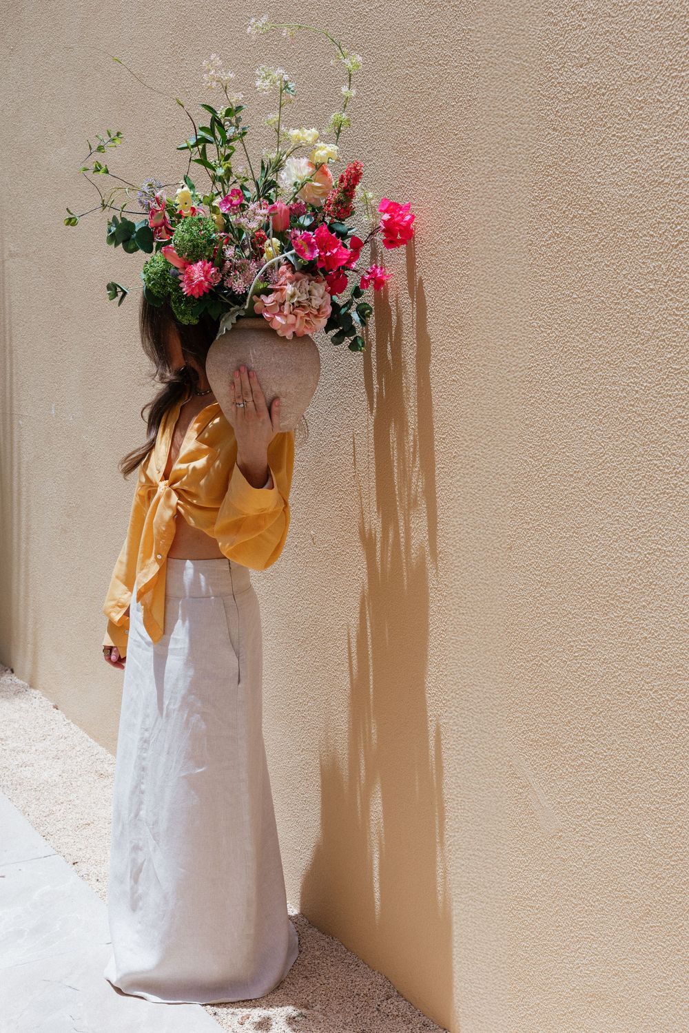 Child in yellow top and white skirt holds large floral arrangement with pink and white blooms against beige wall.