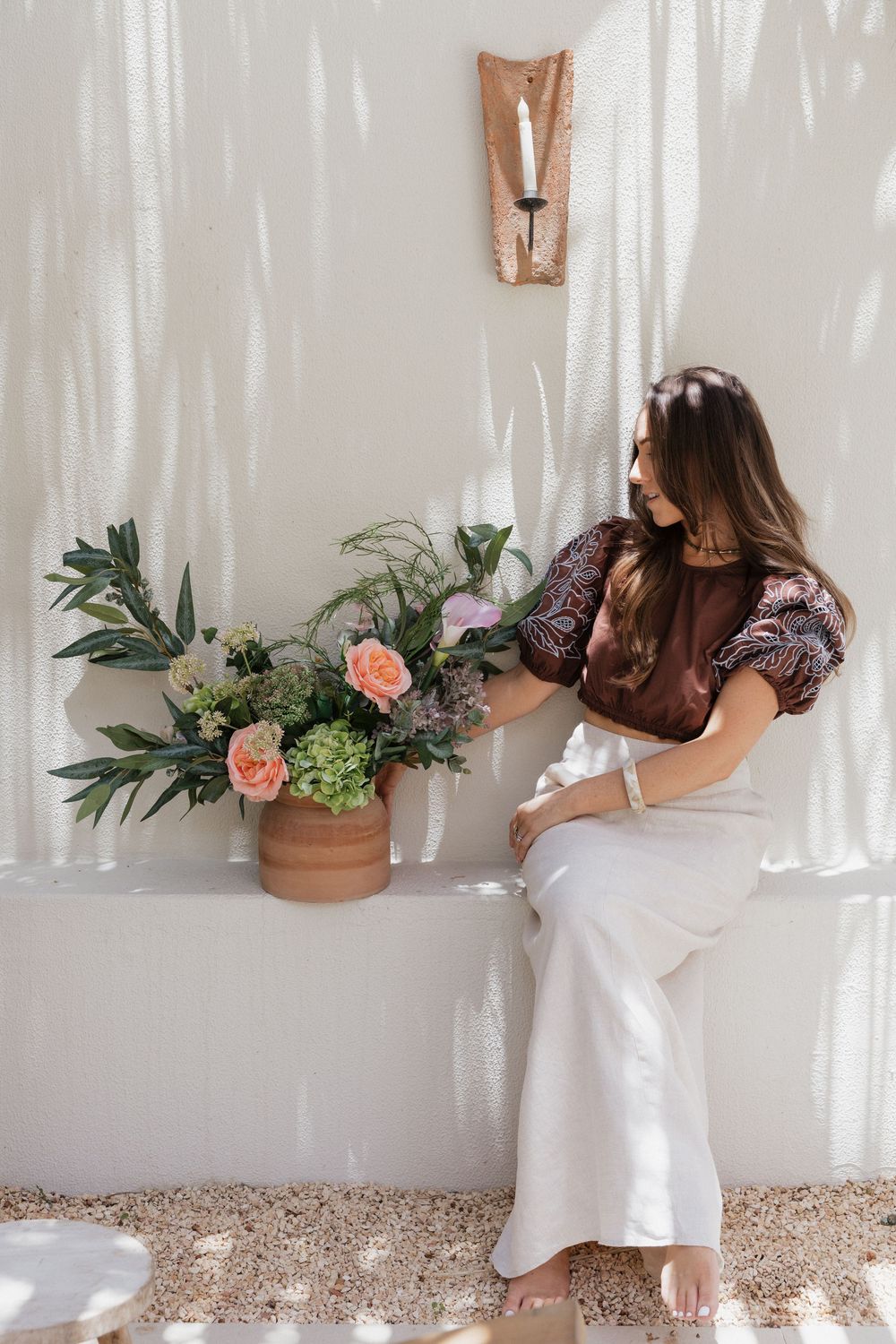 Woman in white pants and floral top sits beside a terracotta pot with pink roses and greenery near a candle sconce.