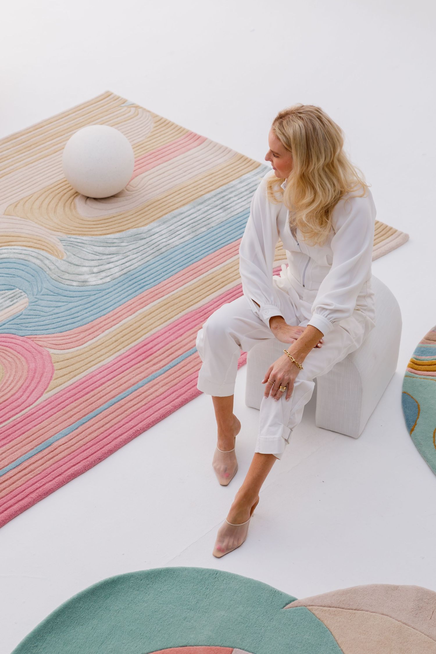 Person in white outfit sits on white surface with colorful wavy patterned rug and sphere decor element in modern minimalist setting.