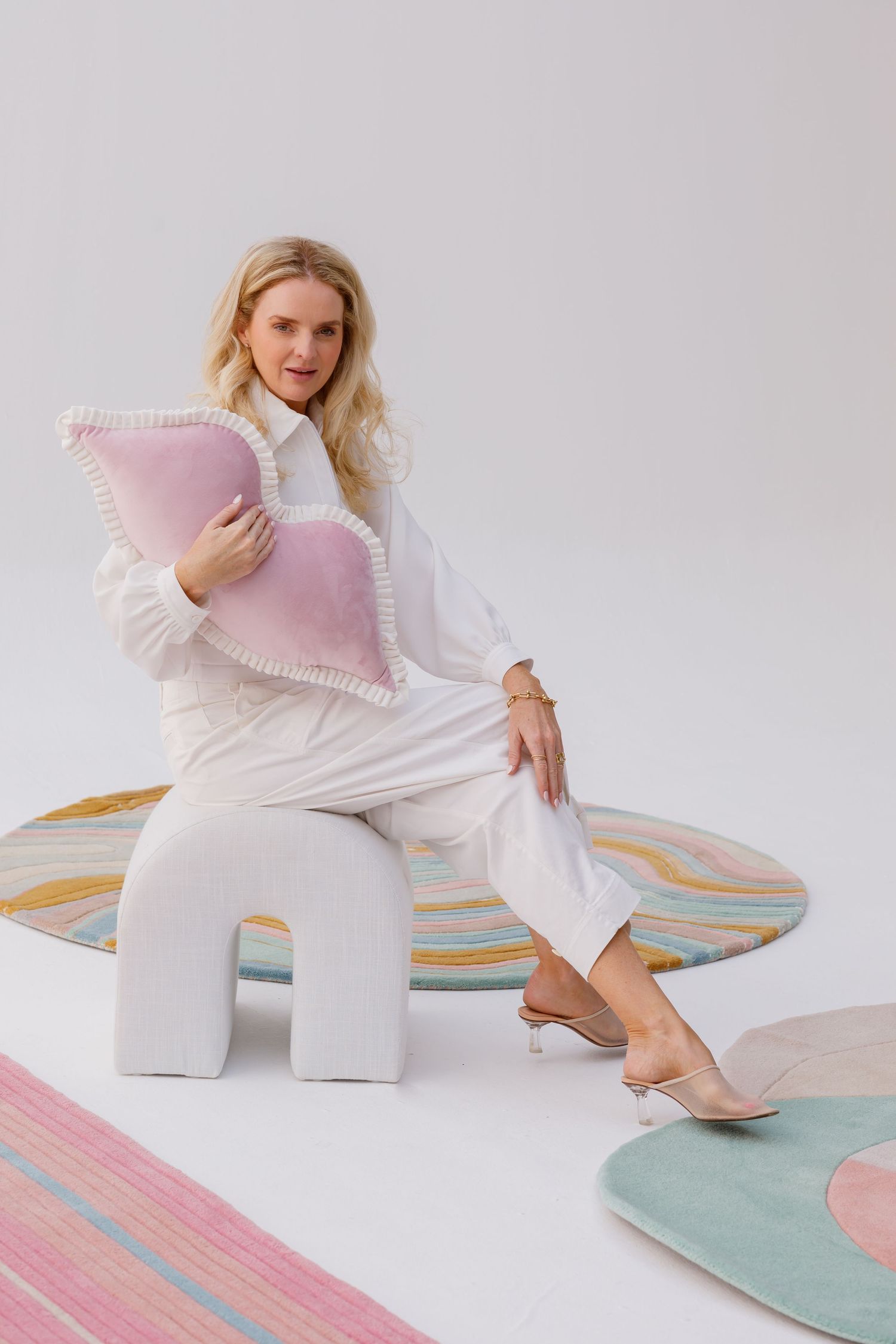 A person in white clothing sits on a modern geometric stool with decorative pastel pillows and rugs in a minimalist setting.