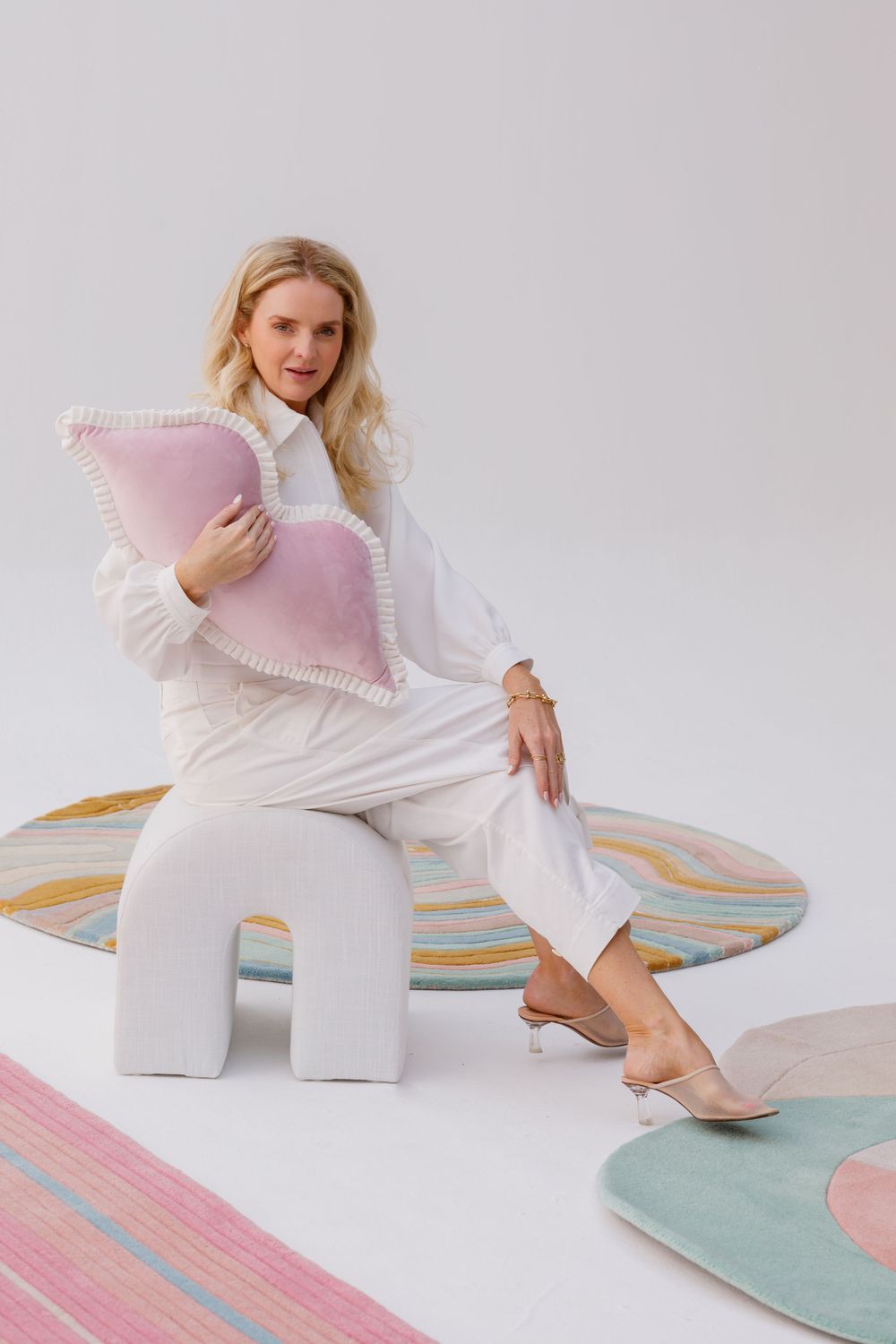 A person in white clothing sits on a modern geometric stool with decorative pastel pillows and rugs in a minimalist setting.