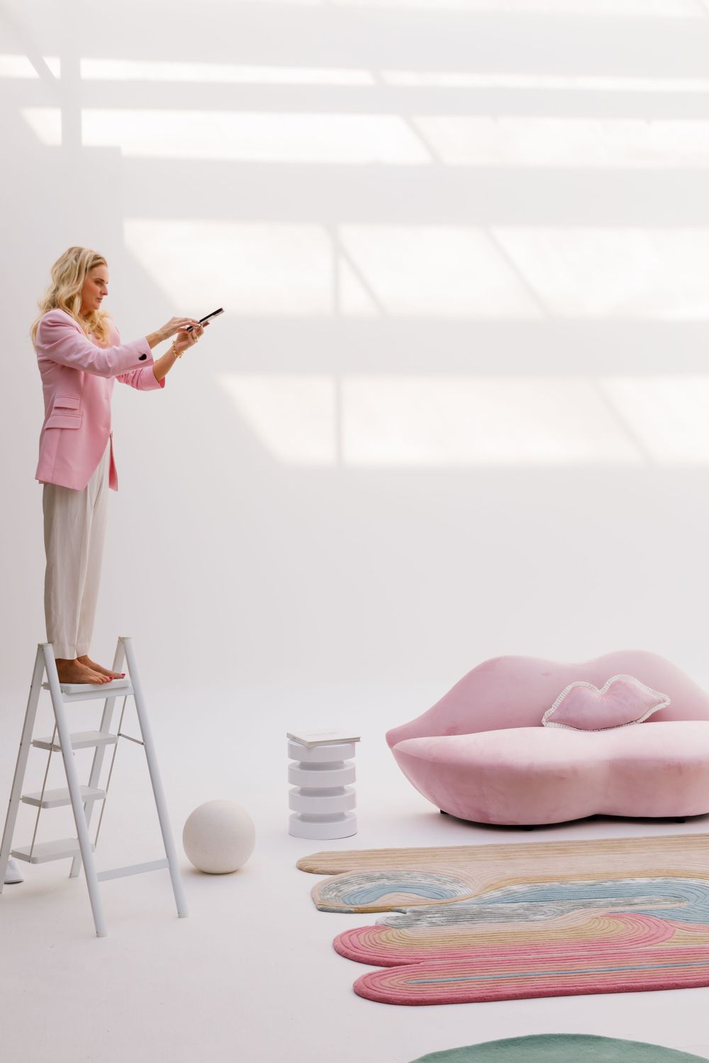 Person in pink blazer stands on ladder in minimalist room with pink sofa and colorful petal-shaped rugs on white floor.