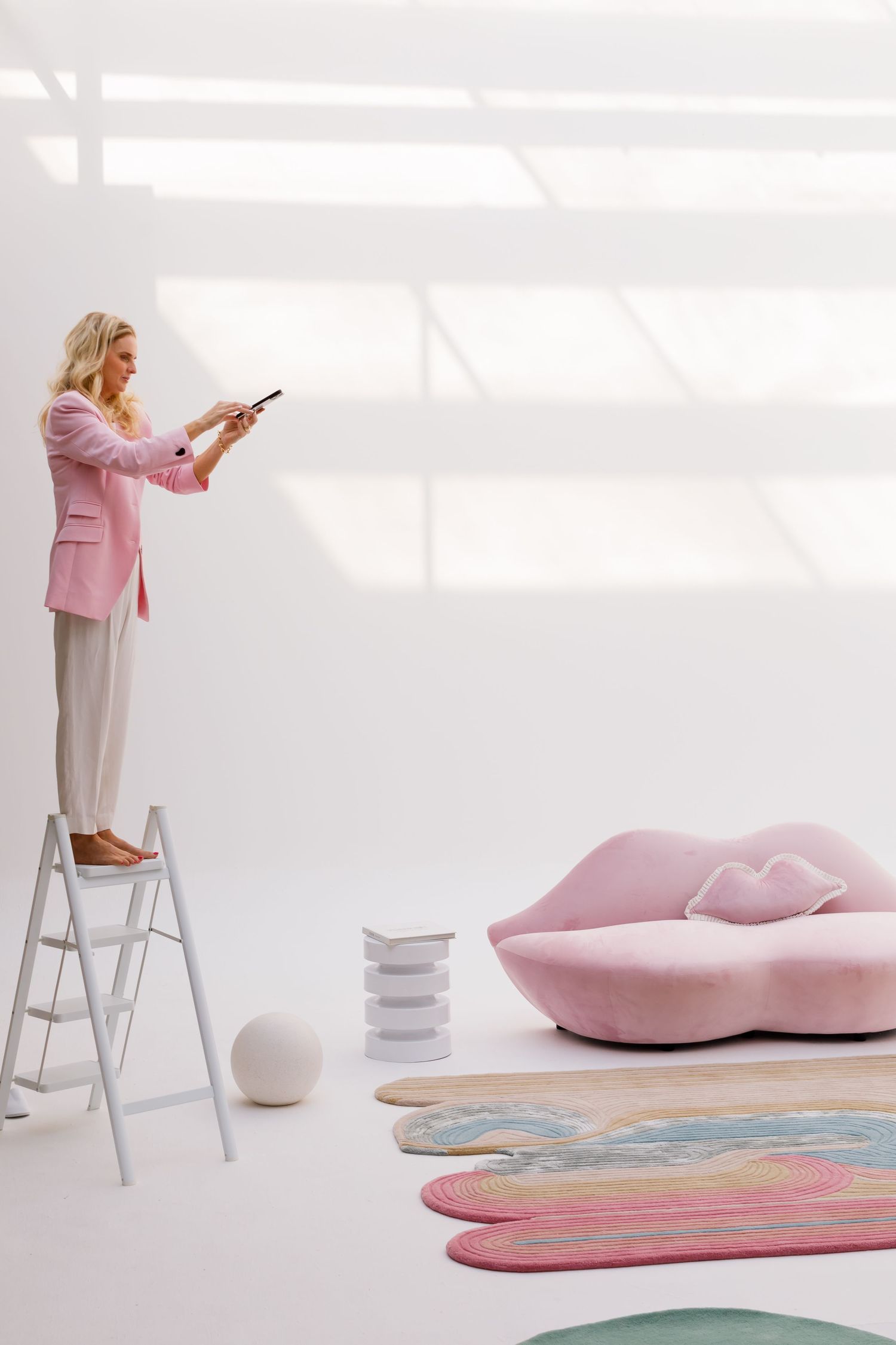 Person in pink blazer stands on ladder in minimalist room with pink sofa and colorful petal-shaped rugs on white floor.